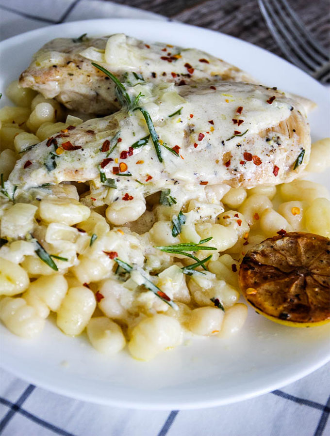 One Pan Creamy lemon chicken is plated with gnocci and fresh herbs on white plate.