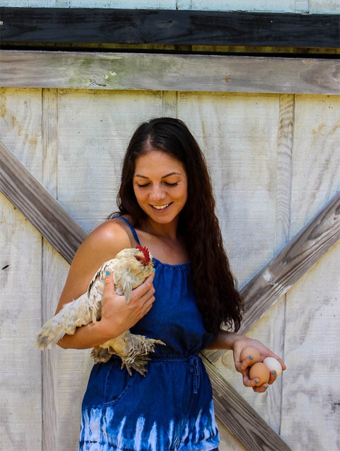 Sherry Brubaker holding fresh eggs and a chicken against a white barn door