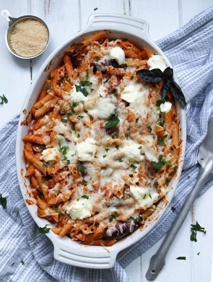 Baked penne pasta is served in a large baking dish next to a bowl of parmesan cheese and a serving spoon.