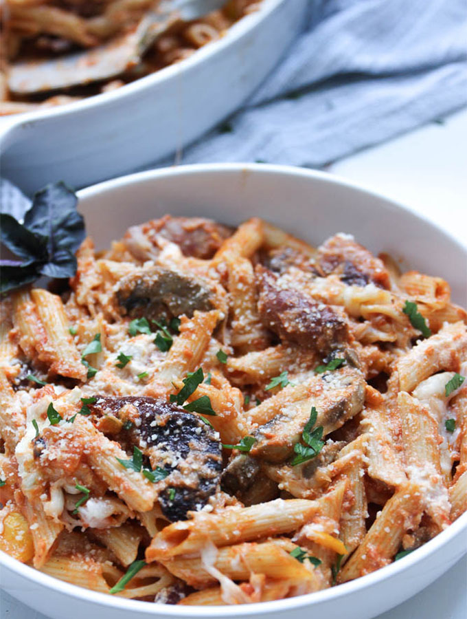 Baked penne pasta is plated in a white bowl and topped with parsley and fresh basil.