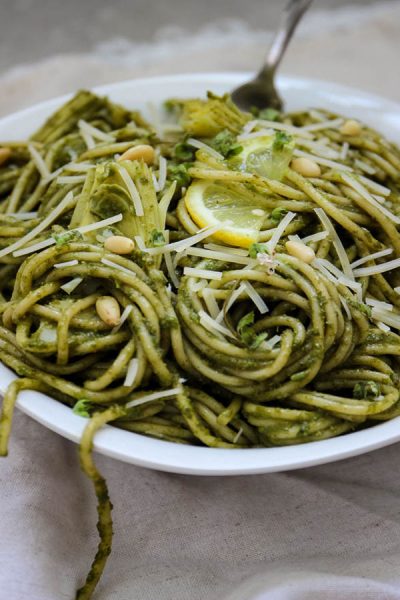 close up shot of pesto pasta with artichoke hearts with a fork and a white bowl