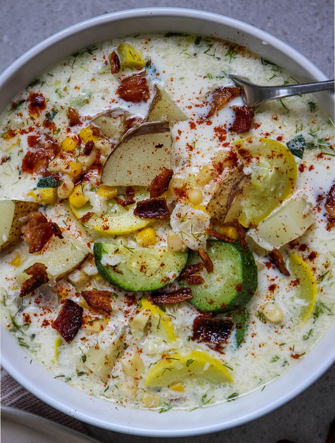 Closeup shot of corn and zucchini chowder to show heaps of colorful zucchini, summer squash, bits of corn, and bite sized pieces of russet potatoes, all topped with crunchy bacon!