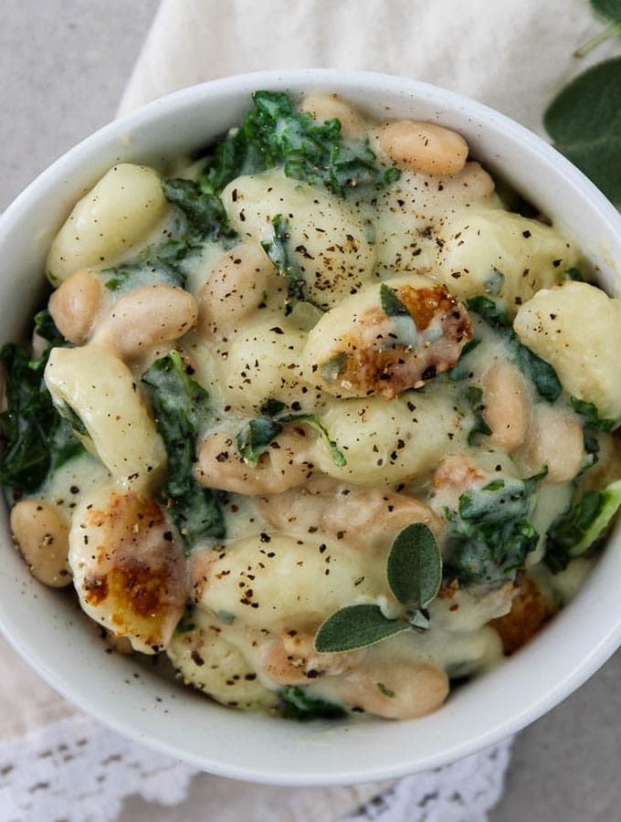 Creamy parmesan gnocchi with kale and white beans is plated in a white dish and topped with cracked pepper.