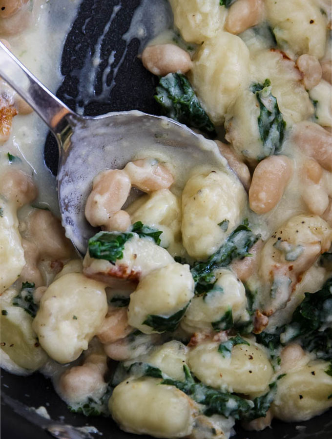 Creamy parmesan gnocchi with kale and white beans is in a non stick pot with a spoon scooping a heaping out.