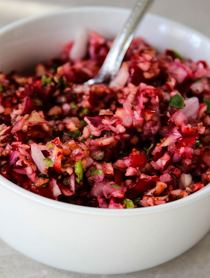 Cranberry relish is plated in a white bowl and is made of cranberries, red onion, serano pepper, oregano, and a splash of balsamic vinegar.