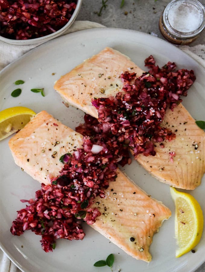 Salmon with Cranberry Relish is plated with heaps of relish on top of the salmon, wedges of lemon for added color, and course salt.