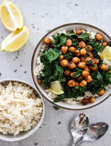 Smoky Chickpea and Kale Rice bowl is plated in a white bowl with a side bowl of rice, a couple of spoons, and wedges of lemon for added color and zest.