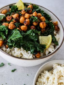 Smoky chickpea and kale rice bowl is plated in a white bowl and topped with small wedges of lemon for added color.