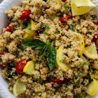 Quinoa salad is plated in a white bowl with a side of lemons and more fresh basil for added color and flavor.
