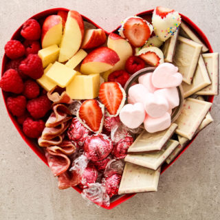 Apples, raspberries strawberries, cheese, salami and valentine's day themed candies are inside of a heart shaped box for a valentine's day charcuterie board.