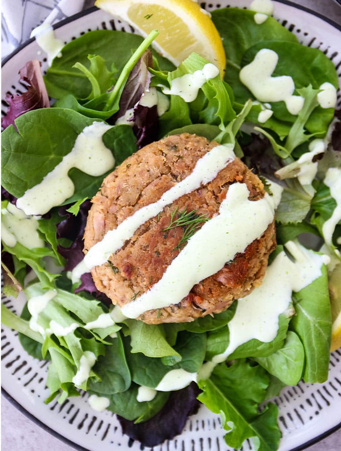 A tuna patty is plated over fresh salad and topped with a lemon wedge and dill sauce.