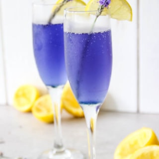 Lavender Lemonade Mimosas are in a flute with fresh lavender and lemon wedges.