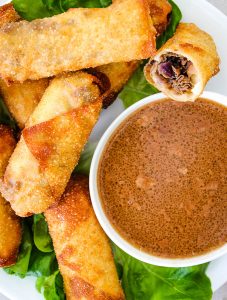 Philly Cheese Steak Egg Rolls are plated around a bowl of au jus sauce.