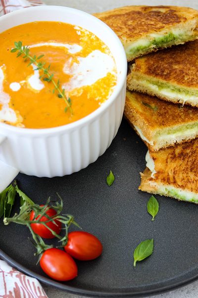 Tomato bisque and grilled cheese sandwiches are plated on a black plate and topped with fresh basil.