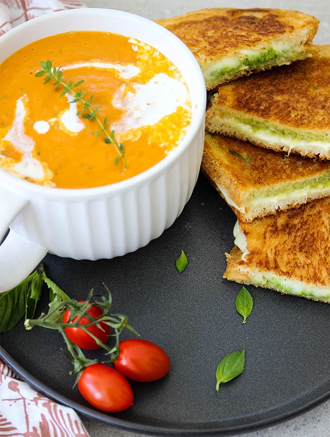 Tomato bisque and grilled cheese sandwiches are plated on a black plate and topped with fresh basil.