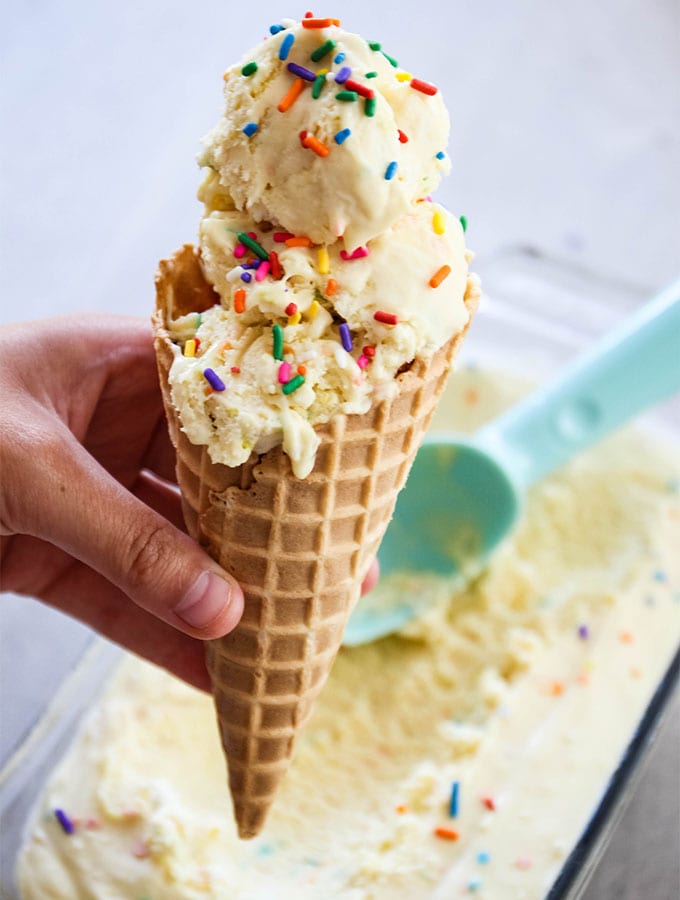 A hand is holding a waffle cone stacked with scoops of No Churn Cake Batter Ice Cream.