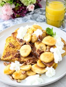 Banana Foster French Toast is plated with orange juice. pecans, and whipped cream.