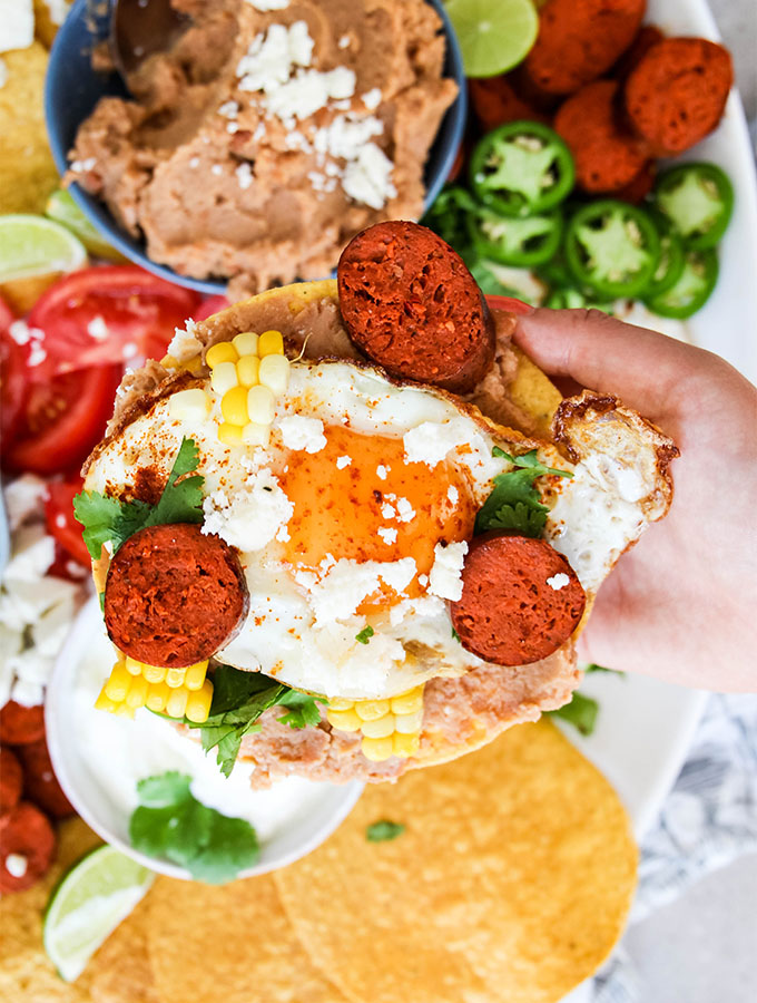 A hand is holding a tostada that is loaded with an egg, sausage, corn, and cheese.