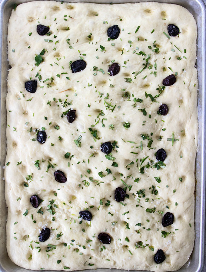 Easy Olive and Herb Focaccia is stretched in a large pan and topped with the fixings.