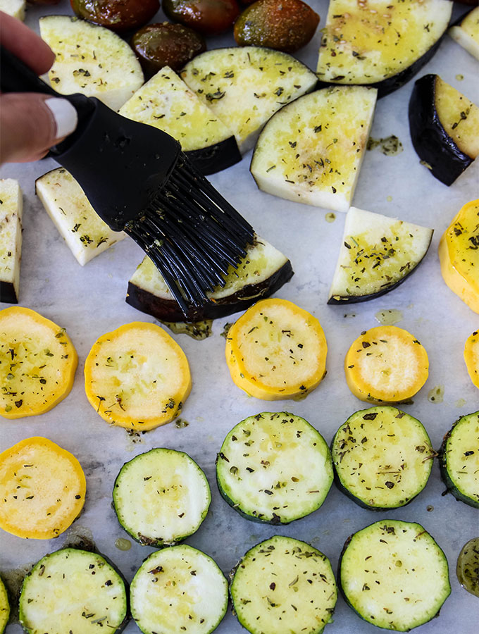 Italian seasoning and olive oil is brushed onto of the roasted vegetables on a baking sheet.