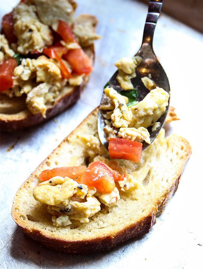 A spoon is placing the scrambled egg bruschetta on a piece of olive oil drizzled toast.