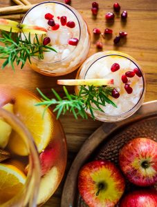 Autumn harvest sangria is poured from the pitcher into glass cups and topped with rosemary and pomegranate arils.