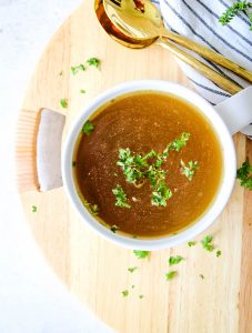 Chicken bone broth is poured into a white soup bowl and topped with parsley and plated next to spoons and a napkin.