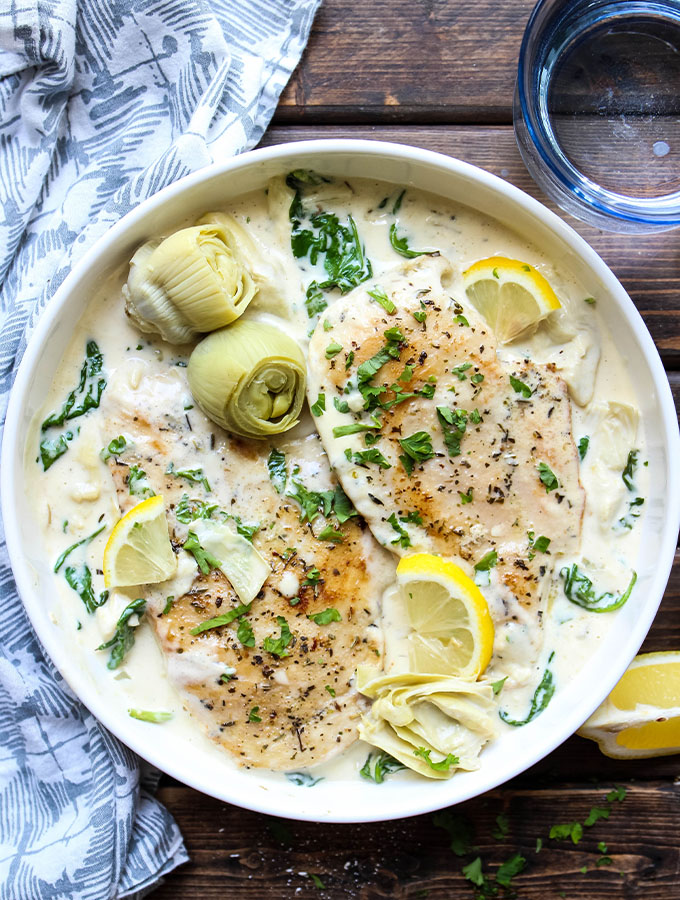 Chicken breasts are plated in a white bowl with the creamy sauce and topped with parsley, artichokes, and lemon wedges.