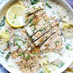 Juicy seared chicken breasts are plated in a white bowl and topped with sauce, cracked pepper, lemons and parsley.