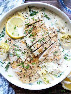 Juicy seared chicken breasts are plated in a white bowl and topped with sauce, cracked pepper, lemons and parsley.