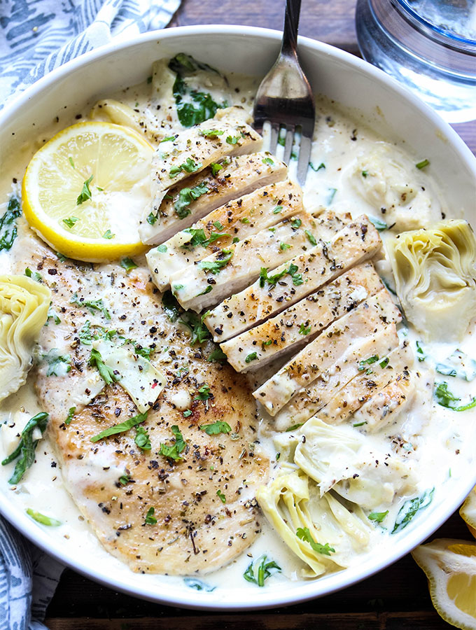 Sliced chicken breasts are plated on top of the herb cream sauce in a white bowl with a fork.