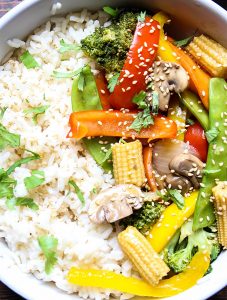 The stir fry vegetables are laid on top of. abed of rice and topped with cilantro and sesame seeds.