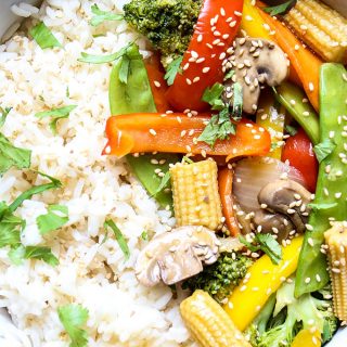 The stir fry vegetables are laid on top of. abed of rice and topped with cilantro and sesame seeds.