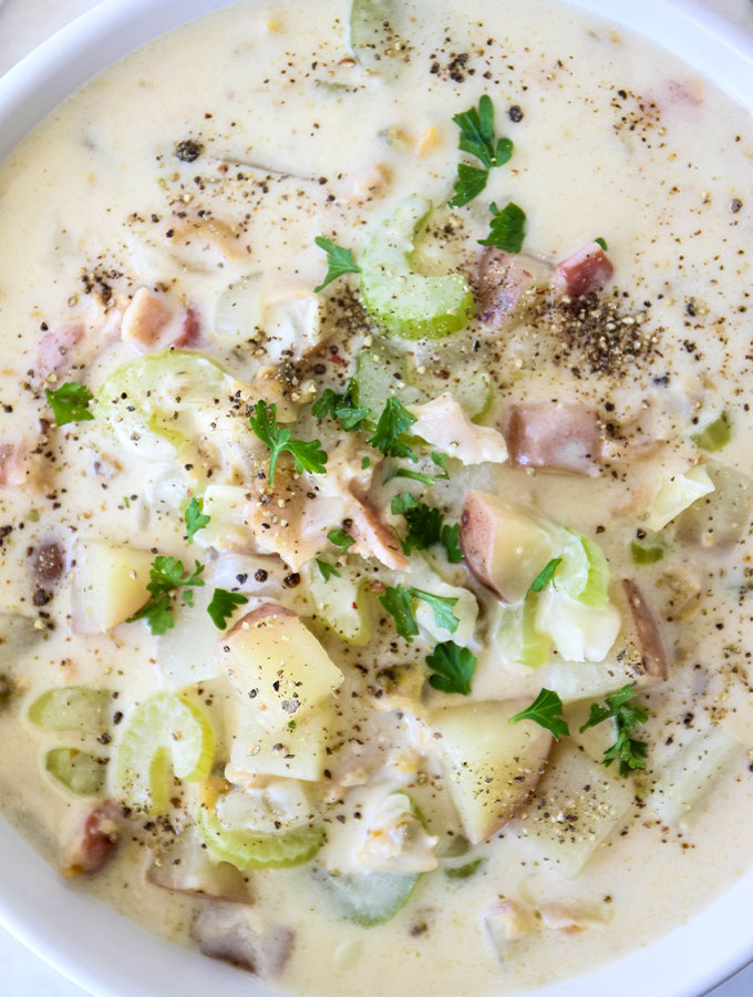 Clam chowder is plated in a white bowl and sprinkled with black pepper and chopped parsley.