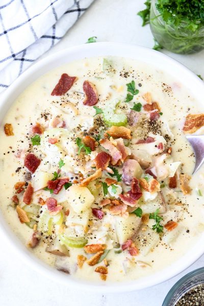 Clam chowder is plated in a white bowl with a spoon and is topped with crispy bacon bits.