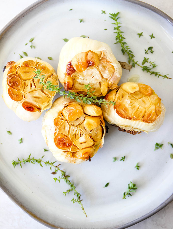 Roasted garlic is plated on a small plate with fresh herbs and a sprinkle of coarse salt.
