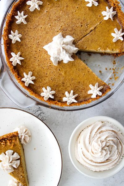 Pumpkin pie is sliced, topped with whipped cream, and a slice is plated on a white plate.