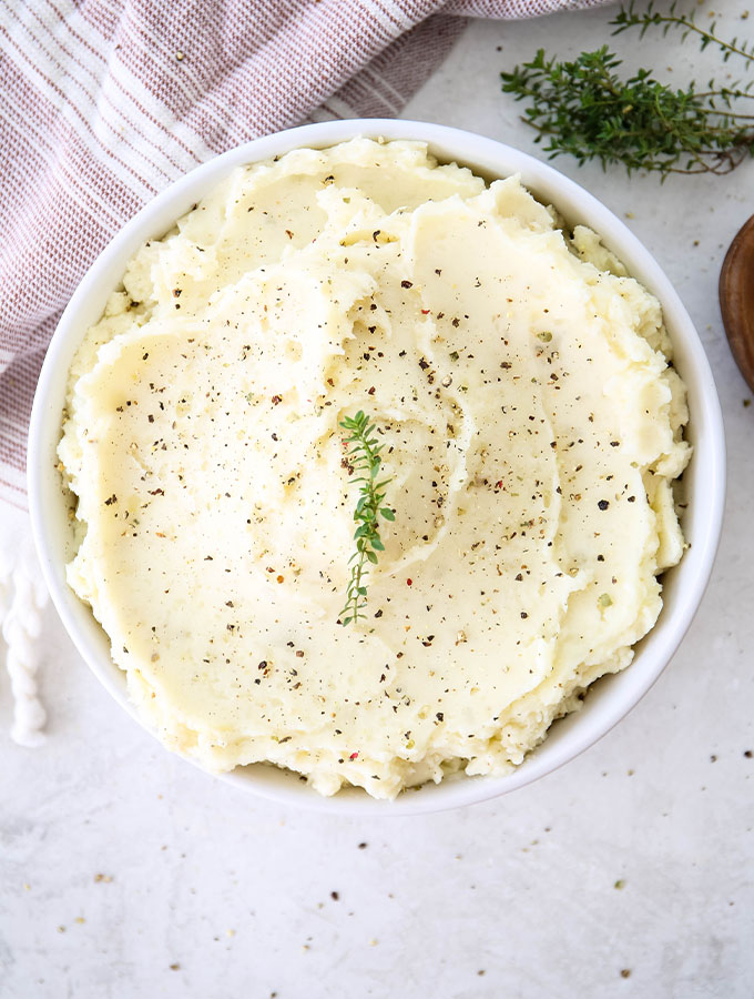 Mashed potatoes are plated in a white bowl and topped with cracked pepper and fresh thyme.