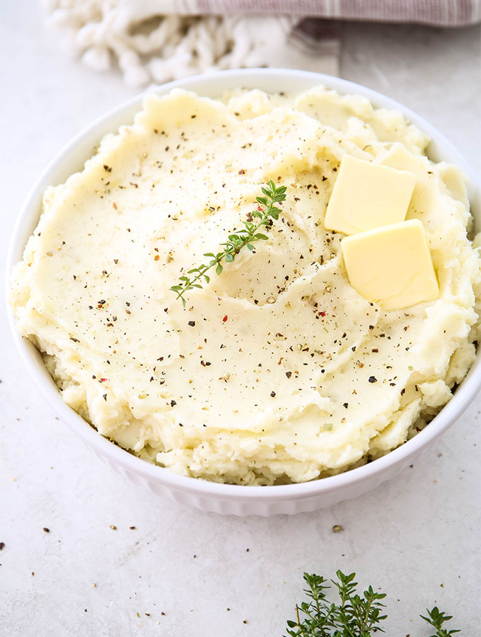 Mashed potatoes are plated in a white bowl and topped with pads of butter, fresh herbs, and cracked pepper.
