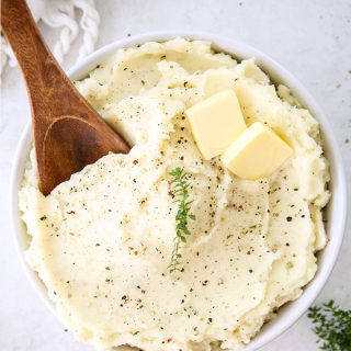 Mashed potatoes are plated in a white bowl with a large wooden spoon spooning some out.