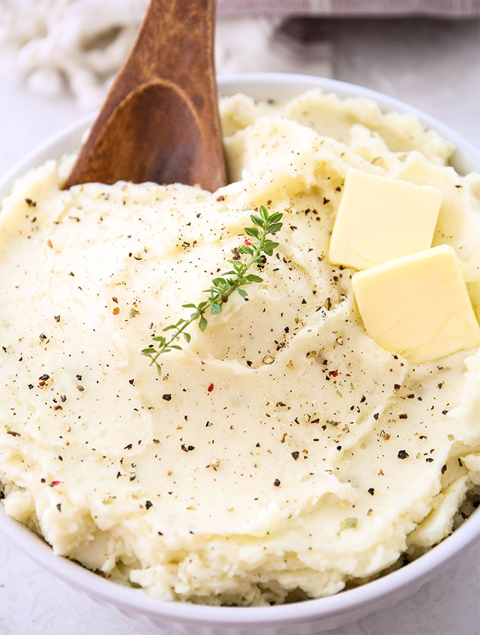 Mashed potatoes are plated in a white bowl with pads of butter, cracked pepper, thyme, and a wooden spoon.