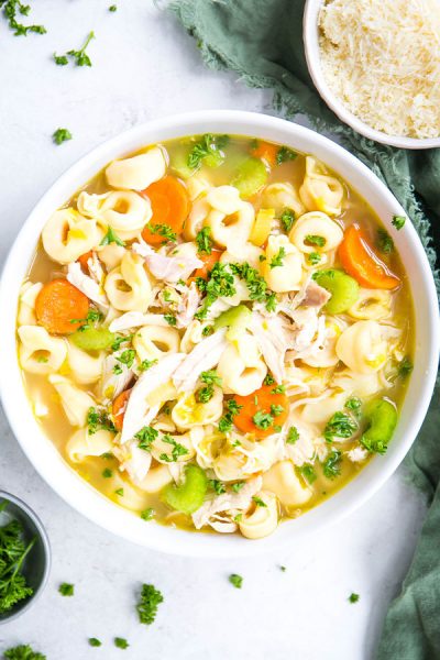 Chicken tortellini is plated in a white bowl and placed next to a bowl of parmesan cheese and fresh parsley.