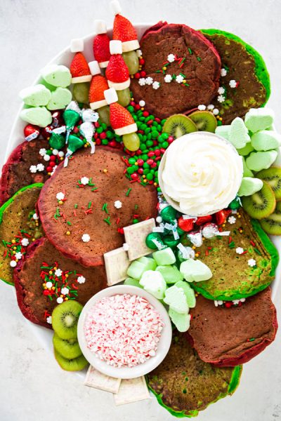 A Christmas breakfast board is filled with pancakes, fruits, candies, and homemade whipped cream.