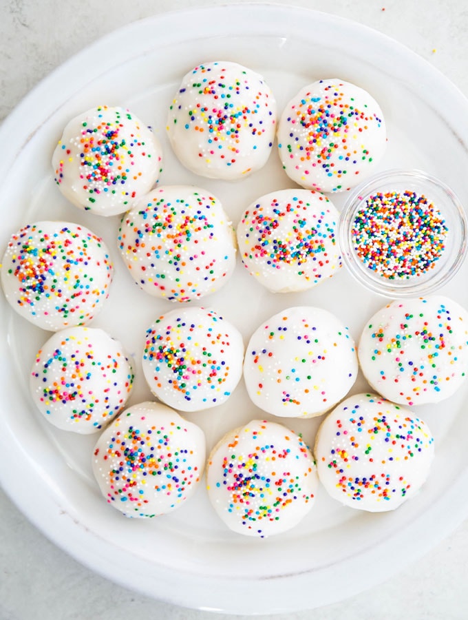Italian anise cookies are plated on a white plate next to a small bowl of sprinkles.