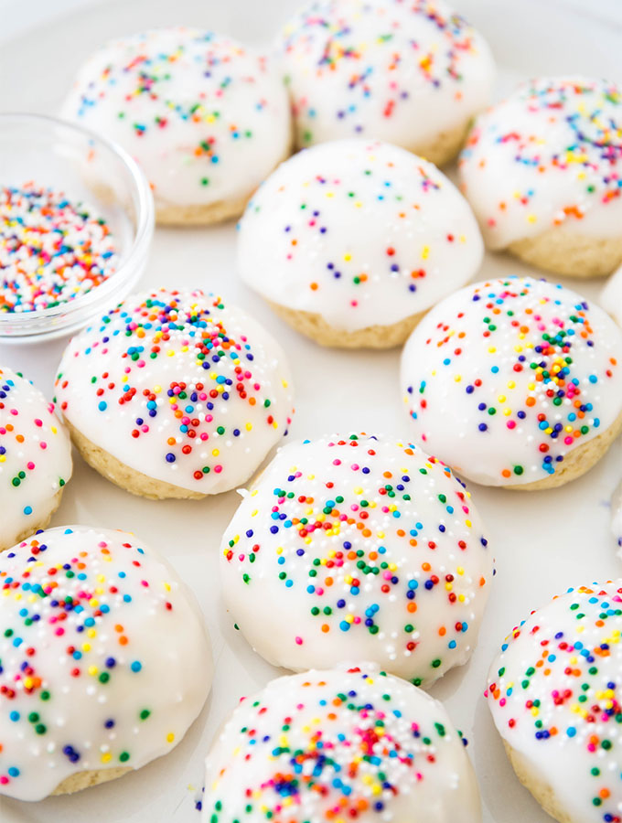Italian cookies are plated on a white plate next to a small bowl of sprinkles.