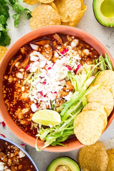 Red pozole is plated in a red bowl and topped with lettuce, radishes, limes, cheese, and chips.