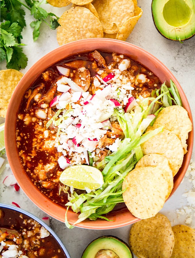 Red pozole is plated in a red bowl and topped with lettuce, radishes, limes, cheese, and chips.