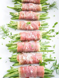 Prosciutto wrapped green beans are plated on a white plate and topped with fresh parsley and parmesan cheese.