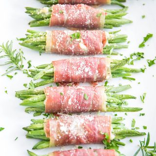 Prosciutto wrapped green beans are plated on a white plate and topped with fresh parsley and parmesan cheese.