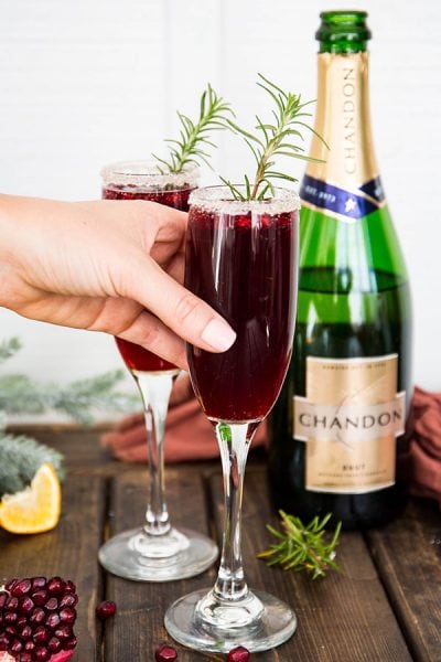 A hand is reaching for a champagne flute that is garnished with rosemary.
